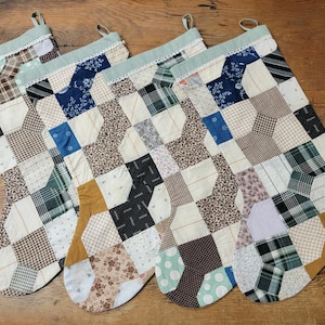May include: Four Christmas stockings with a patchwork design. The stockings feature a variety of fabrics in neutral tones, including beige, brown, and cream, with accents of blue, green, and black. Each stocking has a light green top with lace trim.