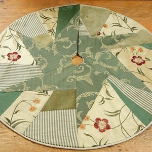 May include: Circular Christmas tree skirt with a patchwork design. The skirt features various fabrics in cream, green, and sage green, with floral and striped patterns. A central opening is surrounded by a decorative border.