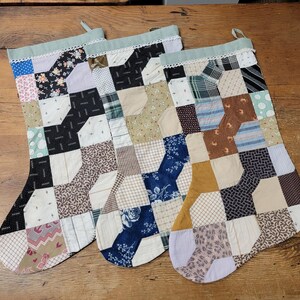 May include: Three handmade Christmas stockings with a patchwork design. Each stocking features a variety of fabric patterns, including floral, polka dot, and geometric prints, in shades of blue, black, brown, and cream. The tops are trimmed with lace and a green fabric.
