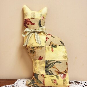 May include: A decorative cat-shaped pillow made from patchwork fabric. The pillow features a floral pattern in yellow, beige, and red hues, with a green and white checkered bow tied around its neck. The cat is sitting on a white lace doily.