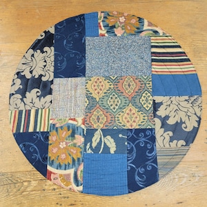 May include: Round patchwork table runner with a variety of blue, brown, and beige fabrics. The fabrics have different patterns, including floral, paisley, and geometric designs.