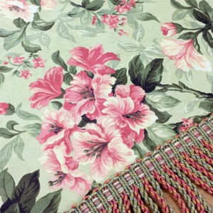 May include: A green fabric with a pink floral pattern and a gold and red fringe trim. The fabric features a large pink flower with a white center.