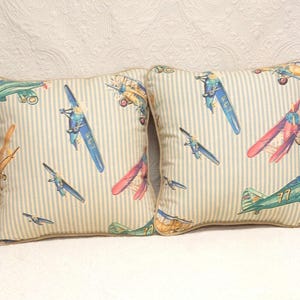 May include: Two decorative throw pillows with a light blue and white striped background. The pillows feature a vintage airplane print in shades of blue, red, green, and yellow.