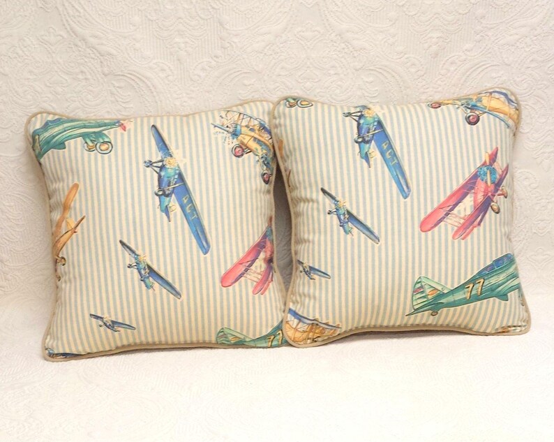 May include: Two decorative throw pillows with a light blue and white striped background. The pillows feature a vintage airplane print in shades of blue, red, green, and yellow.