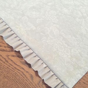 May include: A white table runner with a subtle floral pattern and a ruffled edge. The runner is made of a soft, textured fabric.