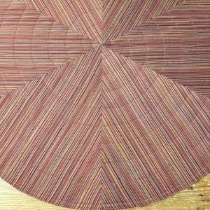 May include: A round, patterned rug with a geometric design. The rug features a central starburst pattern with radiating lines in shades of red, tan, and blue. The edges are curved, and the rug is placed on a wooden surface.