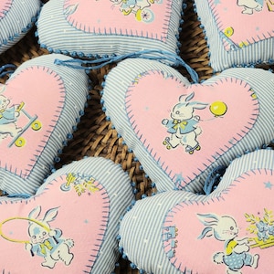 May include: Six pink and blue heart-shaped fabric ornaments with embroidered bunnies and other designs. The ornaments have blue and white stripes and are decorated with white beads.