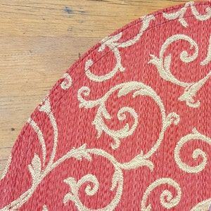 May include: A section of a red fabric with a cream-colored, swirling, floral pattern. The fabric has a textured appearance and is edged with a stitched border. The background is a light brown wooden surface.
