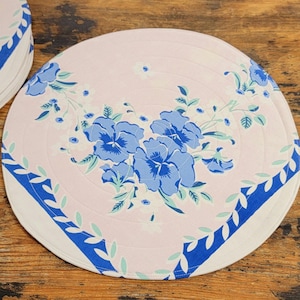 May include: A round pink and blue floral table mat with a blue and white border. The mat features a design of blue pansies and green leaves.