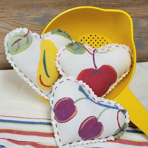 May include: Three heart-shaped fabric cushions with white backgrounds and colourful fruit designs. The cushions are in a yellow colander.