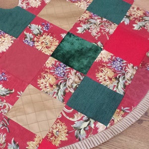 May include: A patchwork Christmas tree skirt in a red, green and gold colour scheme. The skirt is made from various fabrics, including velvet, floral print and plaid. It has a ruffled edge.