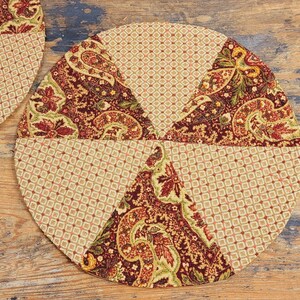 May include: A round, multi-coloured fabric placemat with a paisley pattern. The placemat is made of six fabric panels sewn together. The panels are in shades of brown, beige, and green.