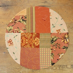 May include: A round, quilted table runner with a patchwork design. The runner is made of various fabrics in shades of red, brown, and cream, featuring floral and geometric patterns.