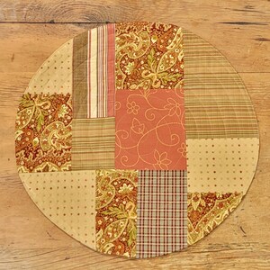 May include: A round, quilted table runner with a patchwork design. The runner is made of various fabrics in shades of brown, beige, and red. The fabrics feature different patterns, including paisley, stripes, and polka dots.