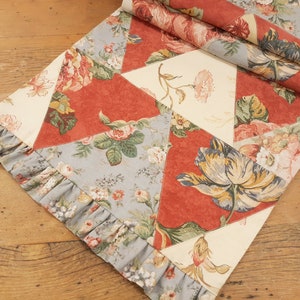 May include: A table runner with a patchwork design featuring a variety of floral patterns in shades of red, blue, and cream. The runner has a ruffled edge and is made of a soft, cotton fabric.