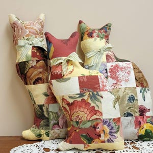 May include: Three decorative cat-shaped pillows. Each pillow is made from patchwork fabric with floral patterns in shades of red, yellow, and green. They are adorned with green and white gingham bows around their necks. The pillows are displayed on a wooden surface.
