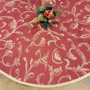 May include: Red Christmas tree skirt with a gold floral pattern and a cream-colored border. The skirt is decorated with a small Christmas tree with ornaments.
