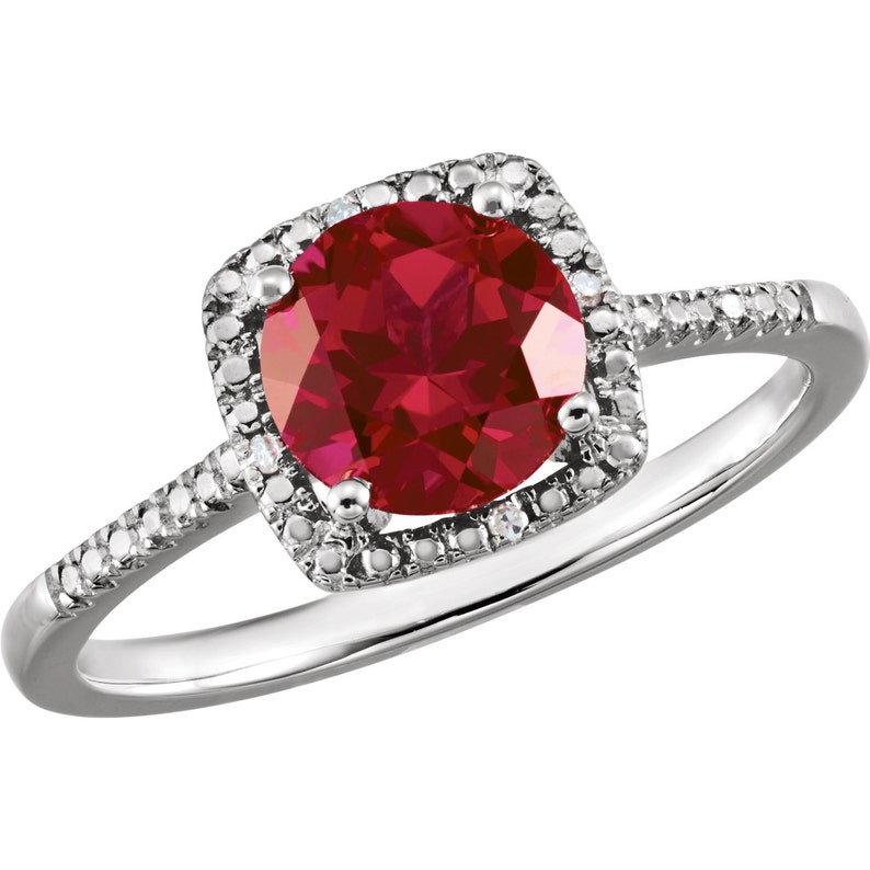 May include: A silver ring with a red gemstone surrounded by a halo of smaller diamonds. The ring has a square-shaped setting and a band that is set with smaller diamonds.