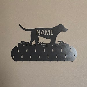 May include: Black metal dog silhouette with the word "NAME" on its back, mounted on a wall with a row of hooks for hanging items.
