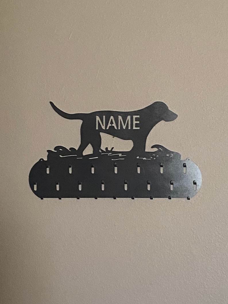 May include: Black metal dog silhouette with the word "NAME" on its back, mounted on a wall with a row of hooks for hanging items.