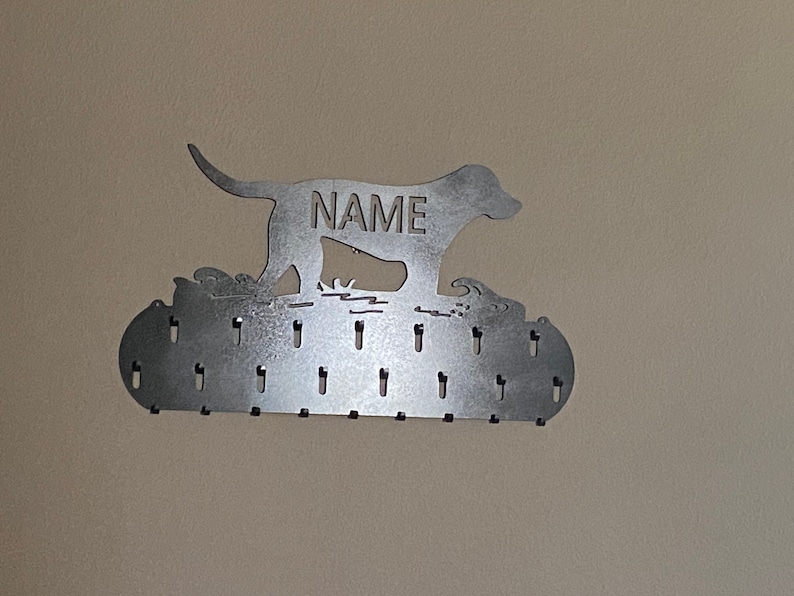 May include: Metal dog silhouette medal holder with the word "NAME" engraved on the dog's body. The holder has 20 hooks for hanging medals.