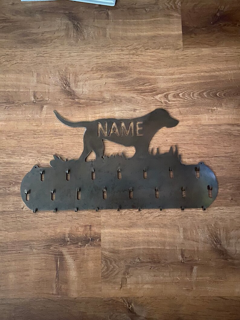 May include: Metal dog silhouette key holder with the word "NAME" engraved on the dog's body. The holder has 20 hooks for hanging keys, leashes, or other items.
