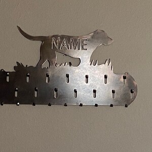 May include: Metal key holder with a silhouette of a dog running, with the word "NAME" in the middle. The holder has 16 hooks for hanging keys.