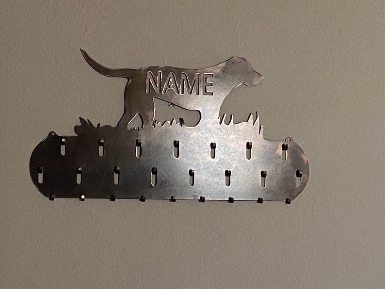 May include: Metal key holder with a silhouette of a dog running, with the word "NAME" in the middle. The holder has 16 hooks for hanging keys.