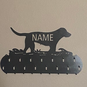May include: Black metal dog silhouette medal holder with the word "NAME" in the center. The holder has 15 hooks for hanging medals.