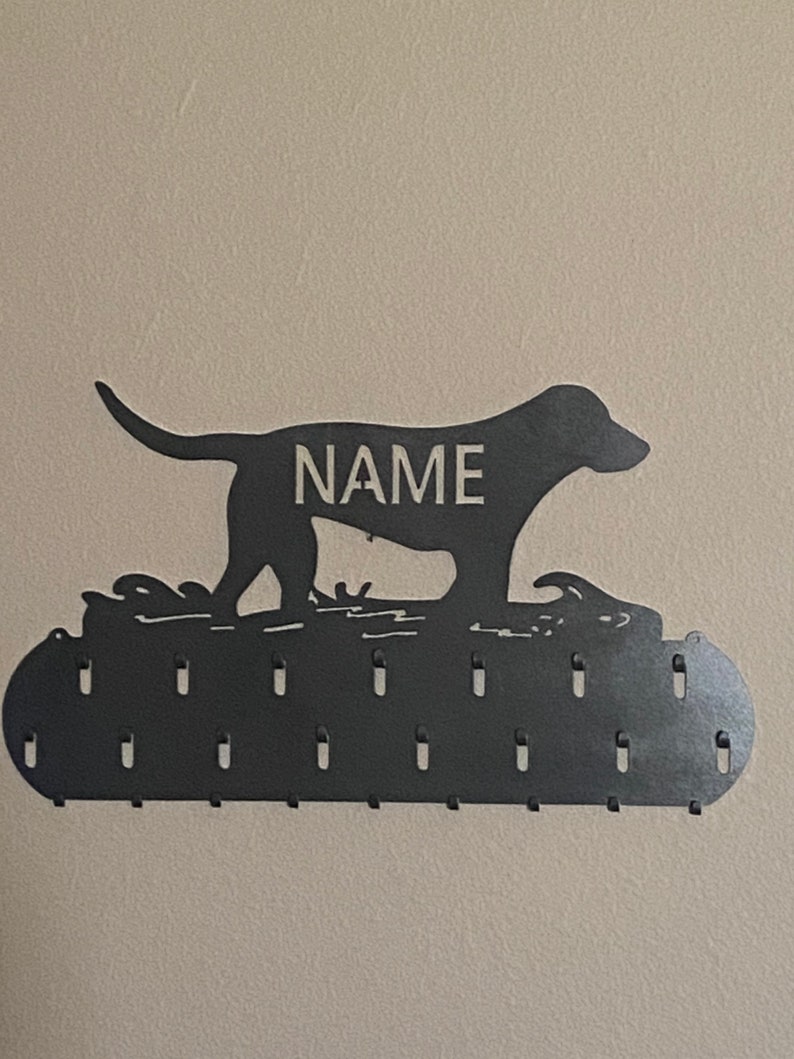 May include: Black metal dog silhouette medal holder with the word "NAME" in the center. The holder has 15 hooks for hanging medals.