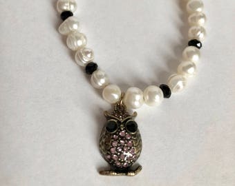 May include: A white pearl bracelet with black beads and an owl charm. The owl charm is made of metal and has pink rhinestones.