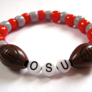 Peut inclure: Un bracelet de perles rouge et gris avec deux breloques de football brunes et des perles de lettres blanches qui épellent "OSU".