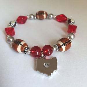 May include: A red and white football themed bracelet with a silver Ohio charm. The charm has a heart cut out of it.