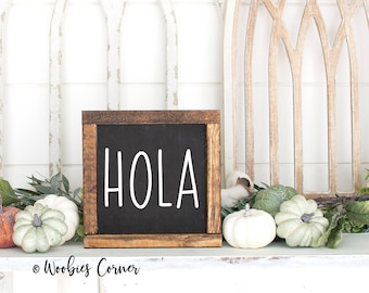 May include: A wooden sign with a black chalkboard insert that says "HOLA" in white lettering. The sign is sitting on a white shelf with a garland of green leaves and white pumpkins.
