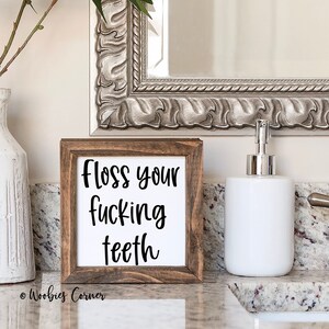 May include: A framed sign with the text "Floss your fucking teeth" in black lettering. The sign is in a brown wooden frame and sits on a bathroom countertop. A white ceramic vase and soap dispenser are also visible.