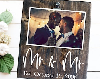 Puede incluir: Un marco de fotos de madera marrón oscuro con un clip blanco en la parte superior. El marco tiene un paspartú blanco y una foto de dos personas con atuendo de boda. El texto "Mr & Mr Est. October 19, 2006" está impreso en blanco en el marco.
