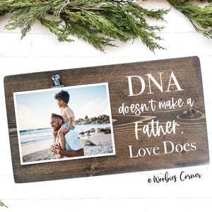 May include: A brown wooden sign with a photo clip holding a photo of a father and son at the beach. The sign reads "DNA doesn't make a Father. Love Does." and is signed "Woobies Corner".