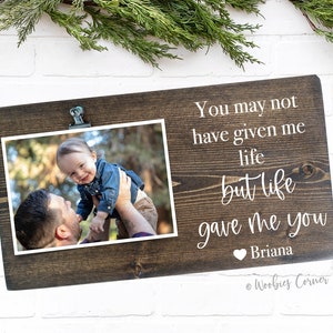 May include: A brown wooden photo frame with a clip at the top. The frame has a white inscription that reads "You may not have given me life but life gave me you Briana". A photo of a man holding a baby is in the frame.