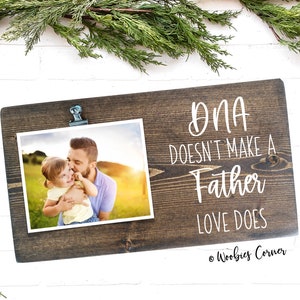 May include: A rustic wooden photo frame with a clip to hold a photo. The frame has the text "DNA doesn't make a father, love does" written on it. The photo shows a father and daughter in a field.