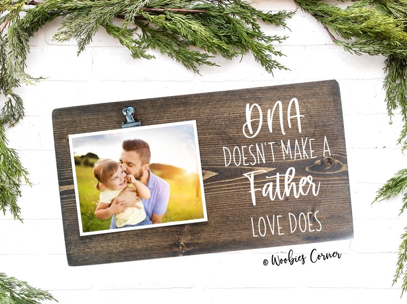 May include: A rustic wooden photo frame with a clip to hold a photo. The frame has the text "DNA doesn't make a father, love does" written on it. The photo shows a father and daughter in a field.