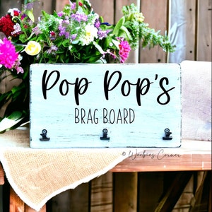 May include: A light blue wooden sign with the text "Pop Pop's Brag Board" in black lettering. The sign has three black metal clips for hanging items.