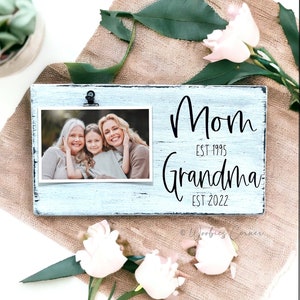 May include: A light blue picture frame with a black border and a clip at the top. The frame has the text "Mom Est 1995 Grandma Est 2022" printed on it. A photo of three women is displayed in the frame.