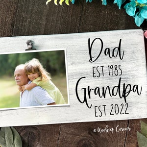 May include: White wooden sign with a photo clip and a photo of an older man and a young girl. The sign says "Dad Est 1985 Grandpa Est 2022" and "Woobies Corner".