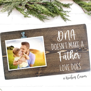 May include: A rustic wooden photo frame with a clip to hold a photo. The frame has the text "DNA doesn't make a father, love does" written on it. The photo shows a father and daughter in a field.