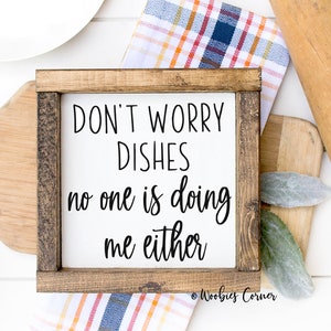 May include: A white wooden sign with a brown frame reads "Don't Worry Dishes no one is doing me either." The sign is sitting on a white surface with a wooden rolling pin, a white plate, and a checkered cloth.