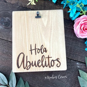 May include: A wooden sign with a clip at the top. The sign is light brown and has the words "Hola Abuelitos" engraved in dark brown. The sign is on a dark brown wooden surface.