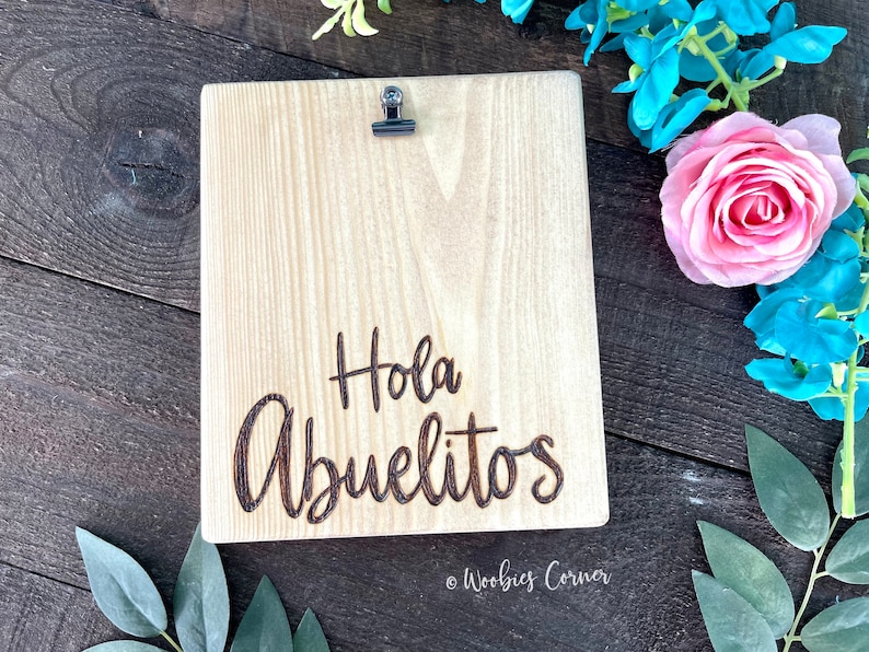May include: A wooden sign with a clip at the top. The sign is light brown and has the words "Hola Abuelitos" engraved in dark brown. The sign is on a dark brown wooden surface.
