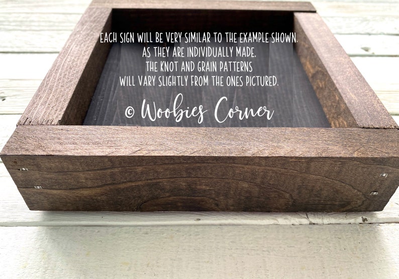 May include: A dark brown wooden frame with a black sign inside. The sign has white text that reads: "EACH SIGN WILL BE VERY SIMILAR TO THE EXAMPLE SHOWN. AS THEY ARE INDIVIDUALLY MADE. THE KNOT AND GRAIN PATTERNS WILL VARY SLIGHTLY FROM THE ONES PICTURED. &copy; Woobies Corner"