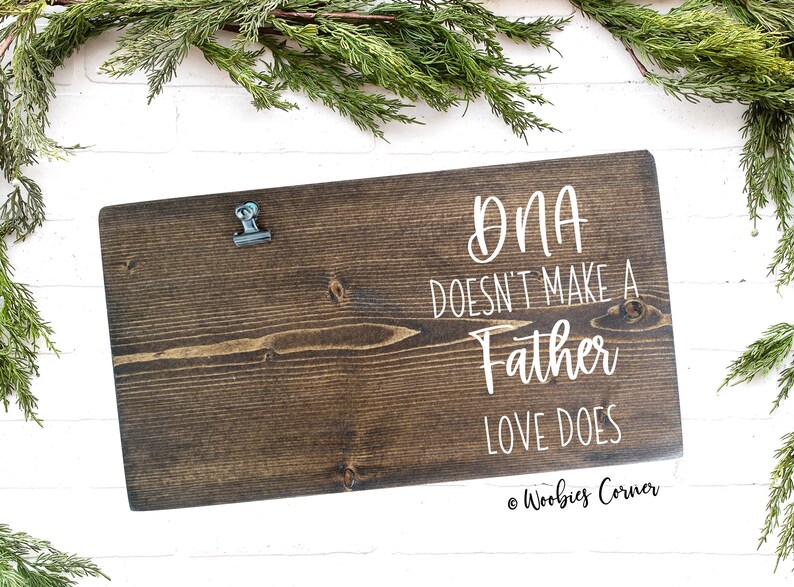 May include: A dark brown wooden sign with a metal clip at the top. The sign has white text that reads "DNA doesn't make a father, love does." The sign is on a white brick background with green foliage.