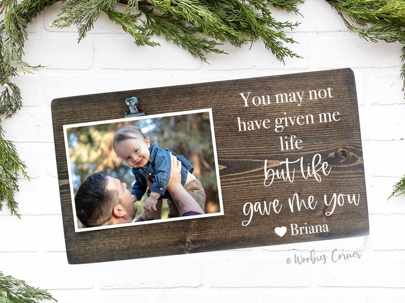May include: A brown wooden photo frame with a clip at the top. The frame has a white inscription that reads "You may not have given me life but life gave me you Briana". A photo of a man holding a baby is in the frame.